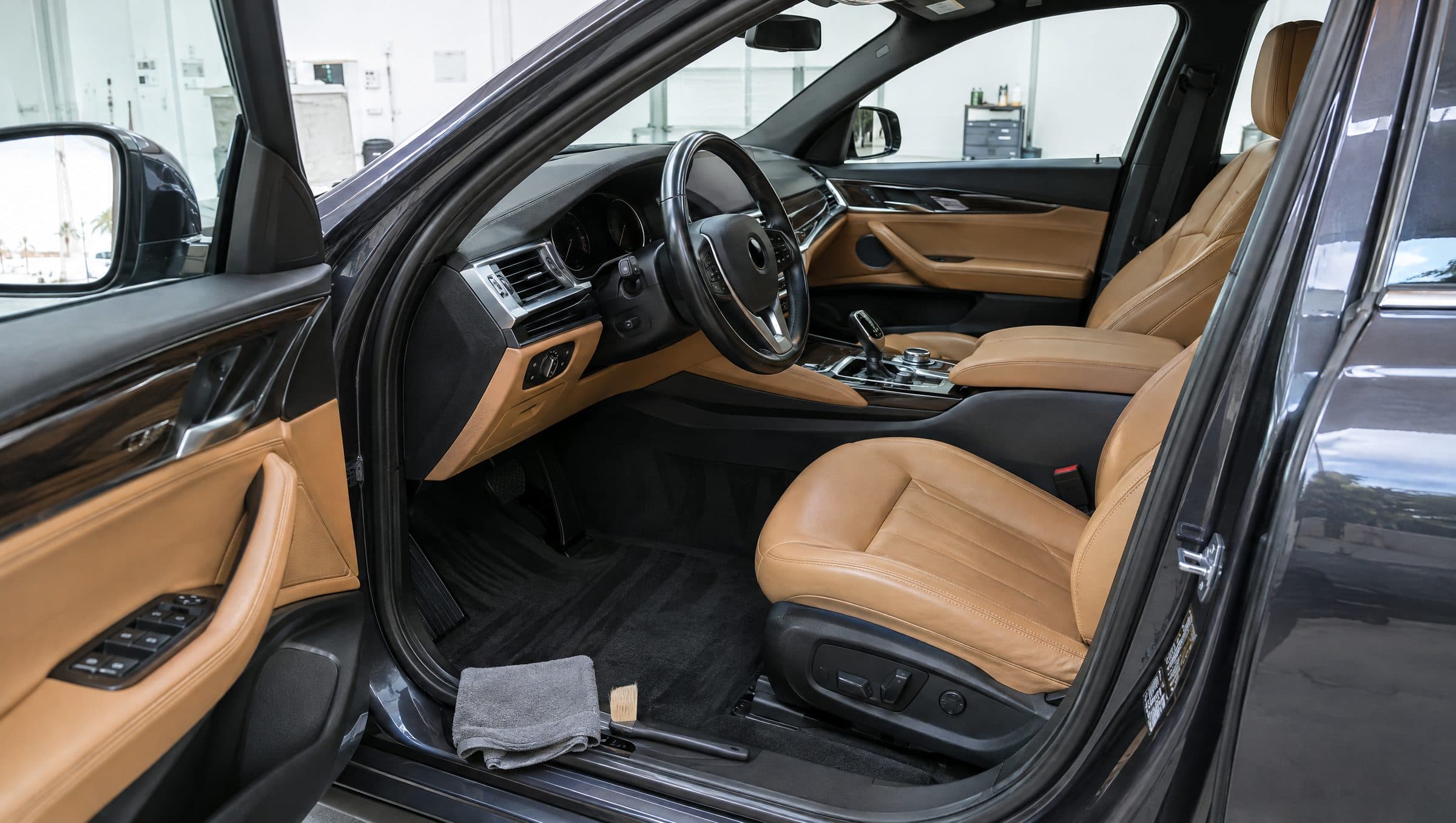 An open driver's door showing a deep-cleaned cabin: tan leather seats, freshly vacuumed black carpets and a clean dashboard.