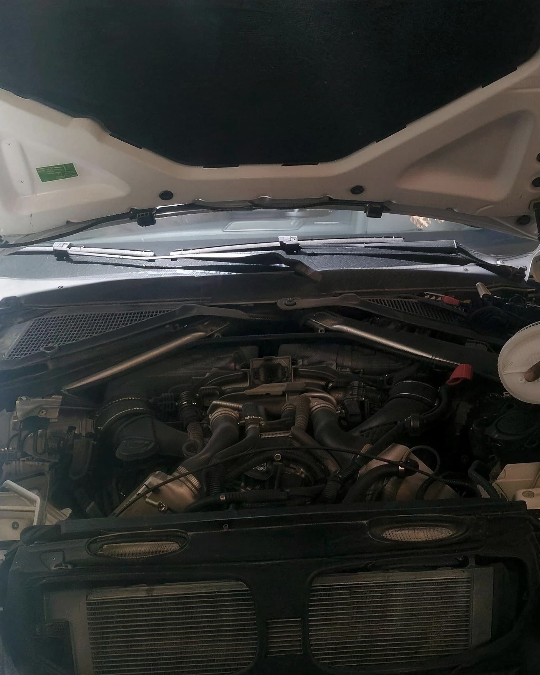 Engine bay of a white BMW X5 with the bonnet up in the workshop, the engine and its pipework exposed above the radiator.