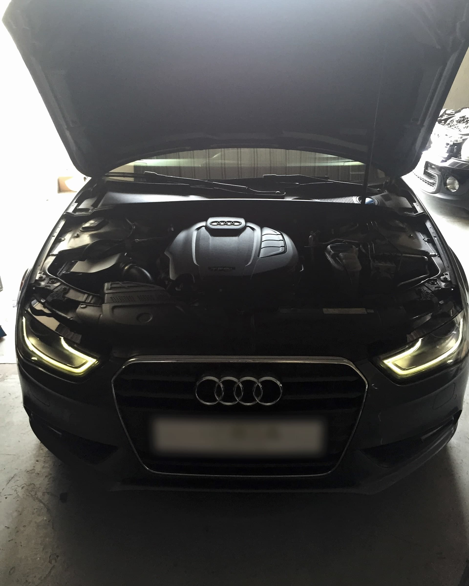 Dark grey Audi A4 in a workshop bay with the bonnet raised, showing the TFSI engine cover, with its daytime running lights on.