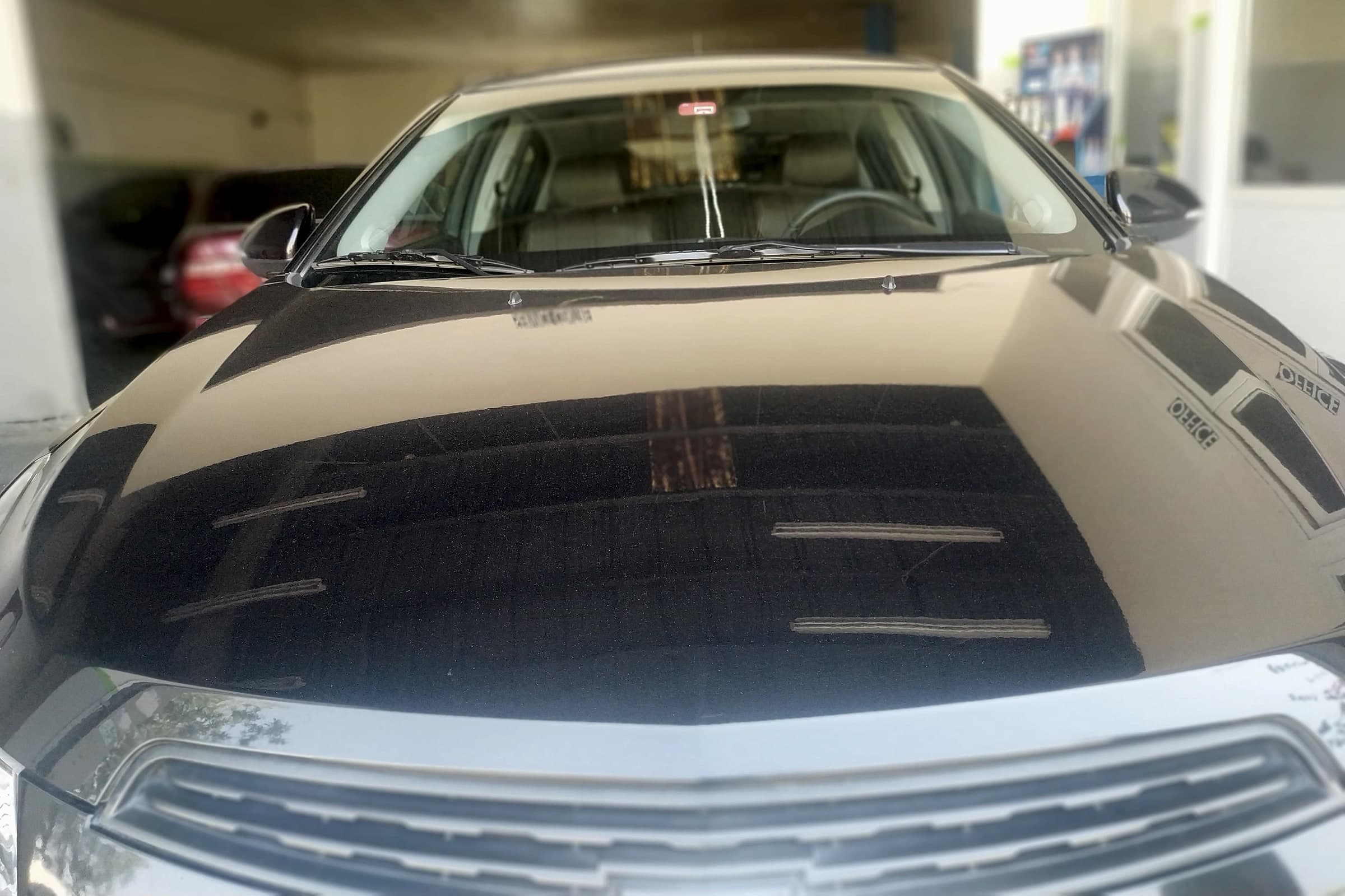 Black Chevrolet parked nose-on in the workshop bay, its glossy bonnet reflecting the ceiling and the office sign.