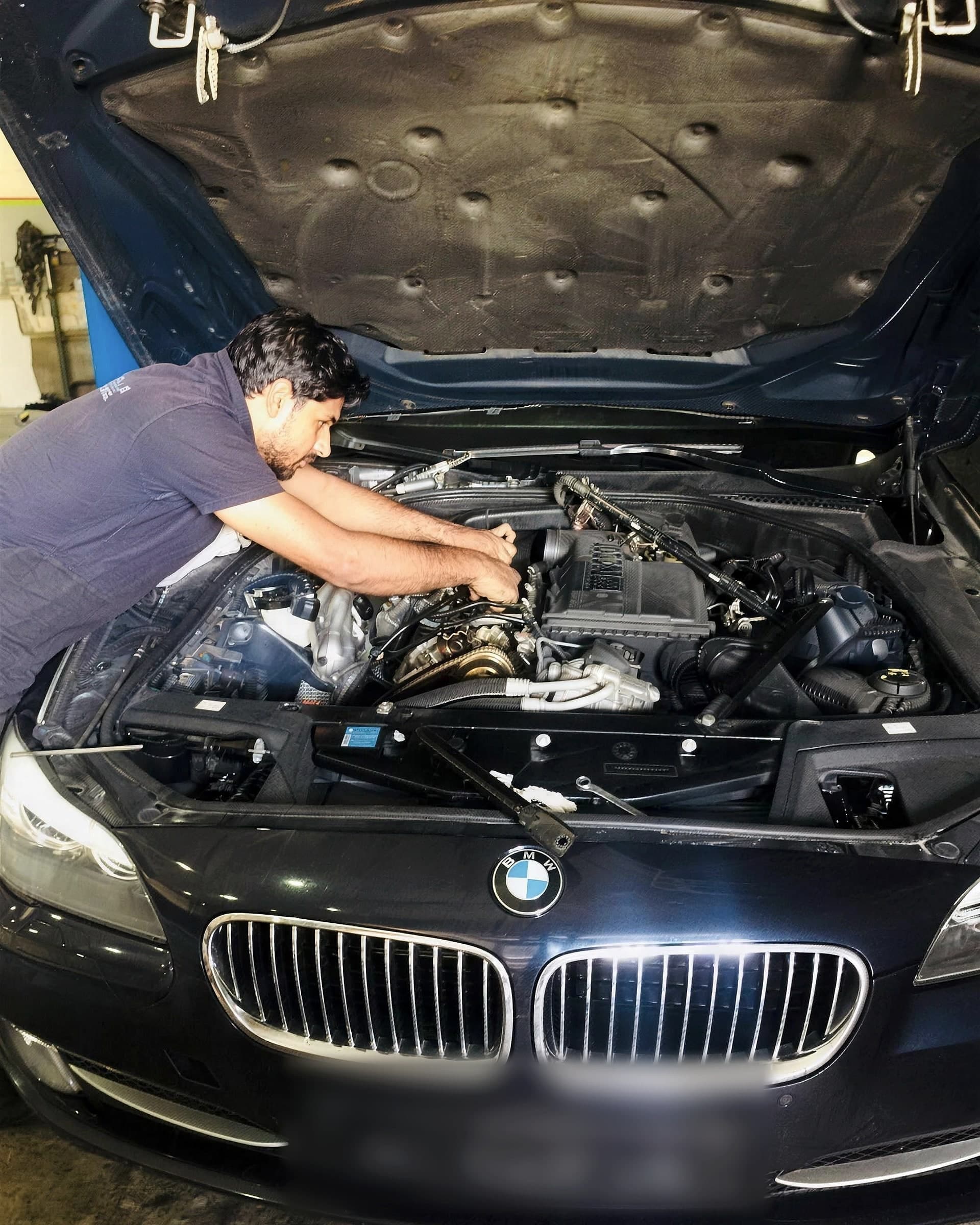 Technician leaning into the engine bay of a dark blue BMW 5 Series with the bonnet up, working on the top of the engine.