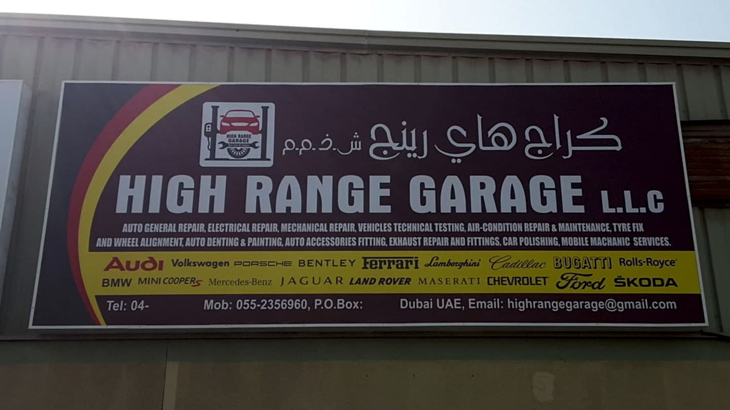 The workshop's maroon sign on its corrugated front wall: High Range Garage L.L.C in English and Arabic, a list of services and a yellow band of the makes it works on.