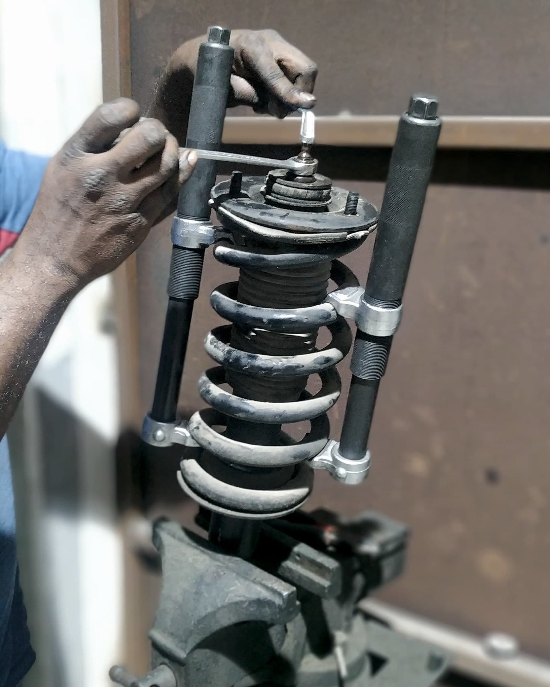 Coil-spring strut held in a spring compressor and clamped in a bench vice while a technician turns the top nut with a spanner.
