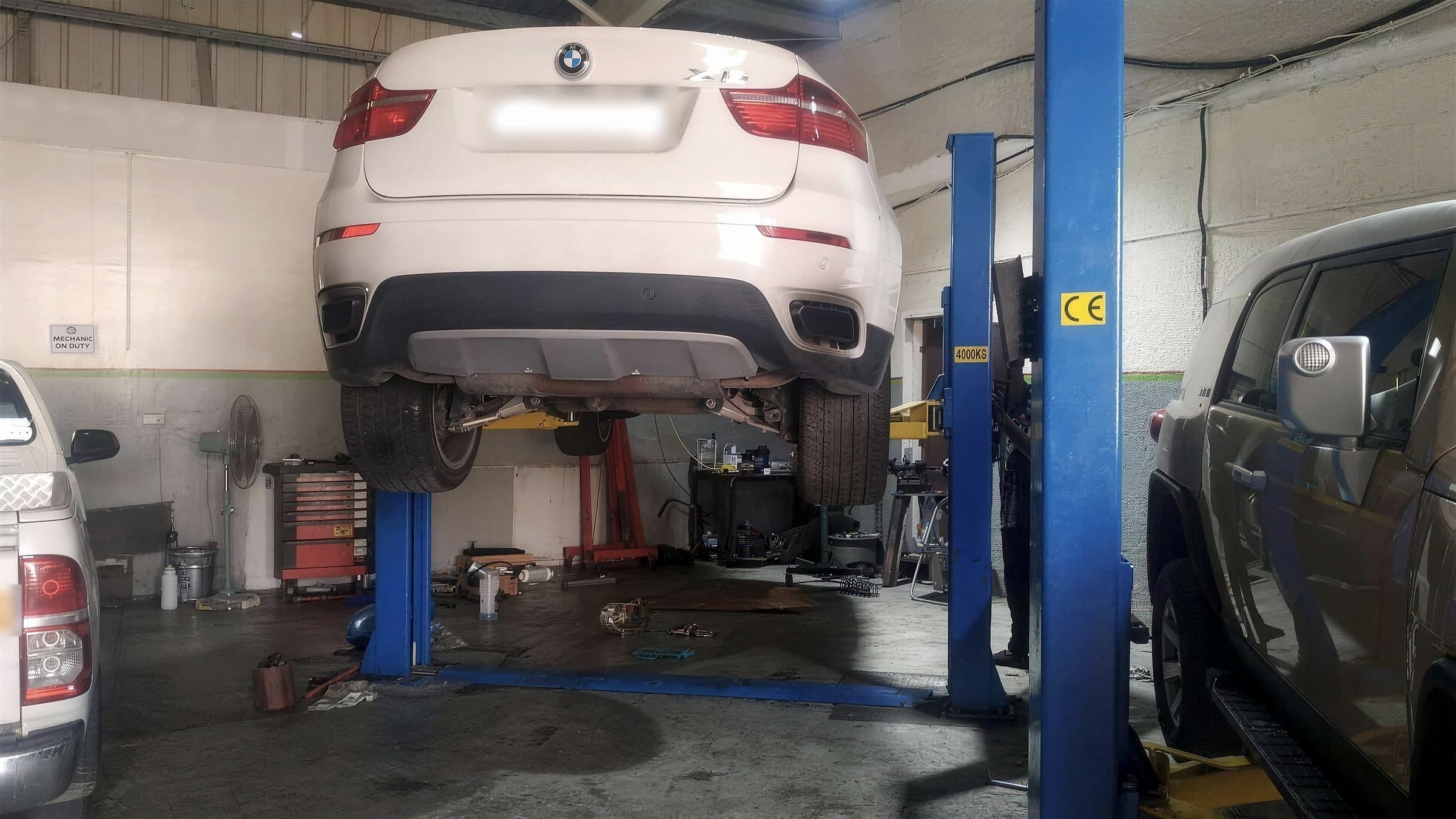 White BMW X6 raised on a blue two-post lift, seen from behind with its wheels hanging and the exhaust and rear suspension exposed, between two other cars in the workshop bays.